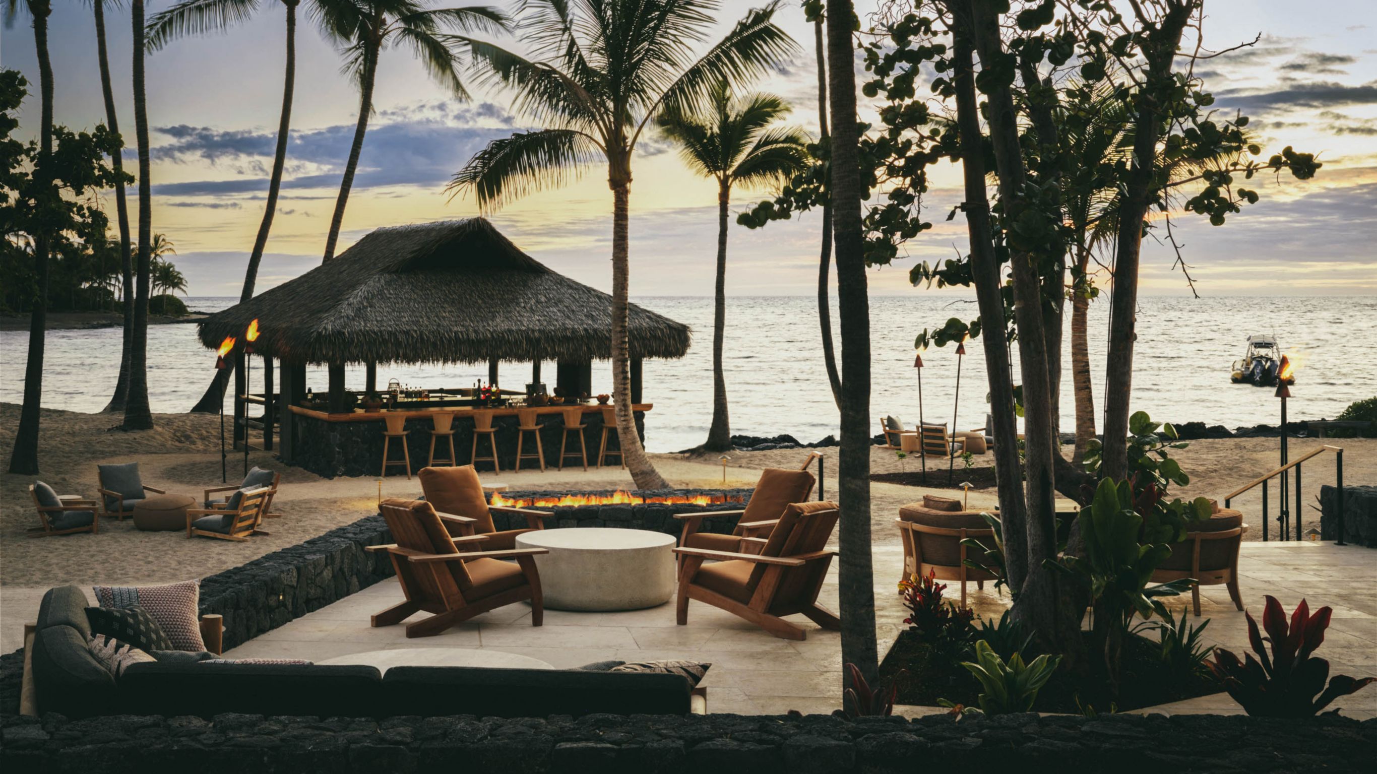 A beachfront area with a thatched roof bar, chairs, a fire pit, and palm trees during sunset.